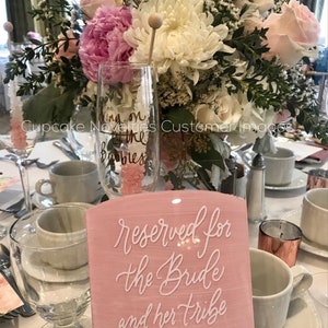 May include: Pink acrylic sign with white calligraphy that reads "reserved for the bride and her tribe". The sign is on a table with a champagne flute and pink flowers.