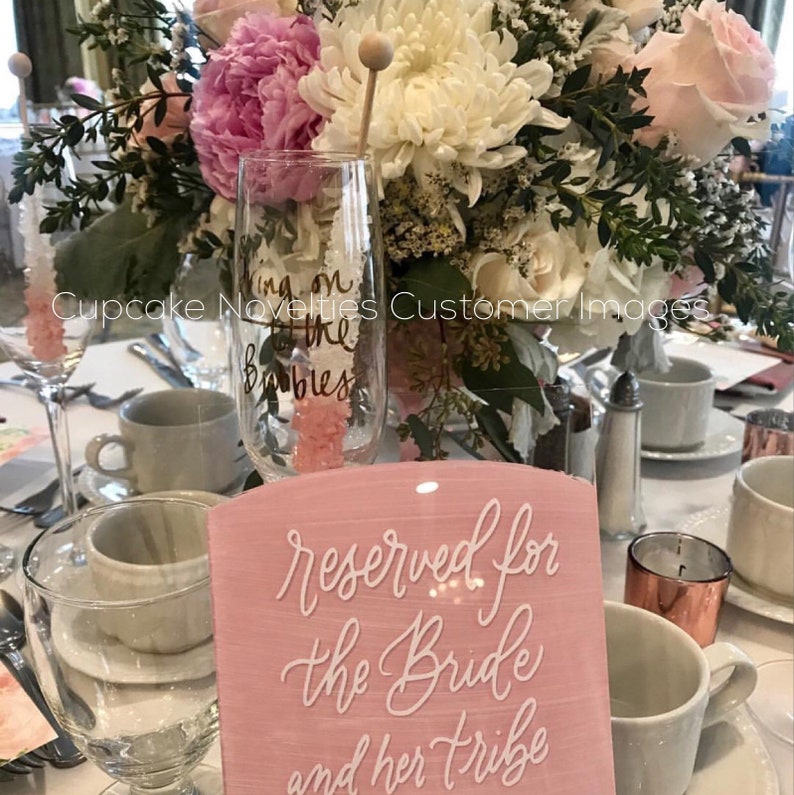 May include: Pink acrylic sign with white calligraphy that reads "reserved for the bride and her tribe". The sign is on a table with a champagne flute and pink flowers.