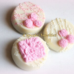 May include: Three white chocolate covered Oreos decorated with pink floral designs and white sprinkles.  The designs include a single rose, a cluster of small flowers, and a larger cluster of flowers.