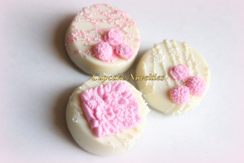 May include: Three white chocolate covered Oreos decorated with pink floral designs and white sprinkles.  The designs include a single rose, a cluster of small flowers, and a larger cluster of flowers.