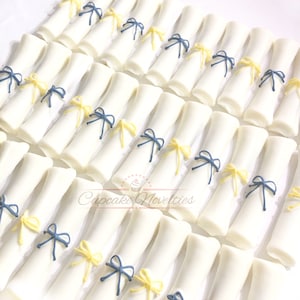 May include: A collection of white fondant graduation caps decorated with yellow and blue ribbon bows. The caps are arranged in rows on a white surface.