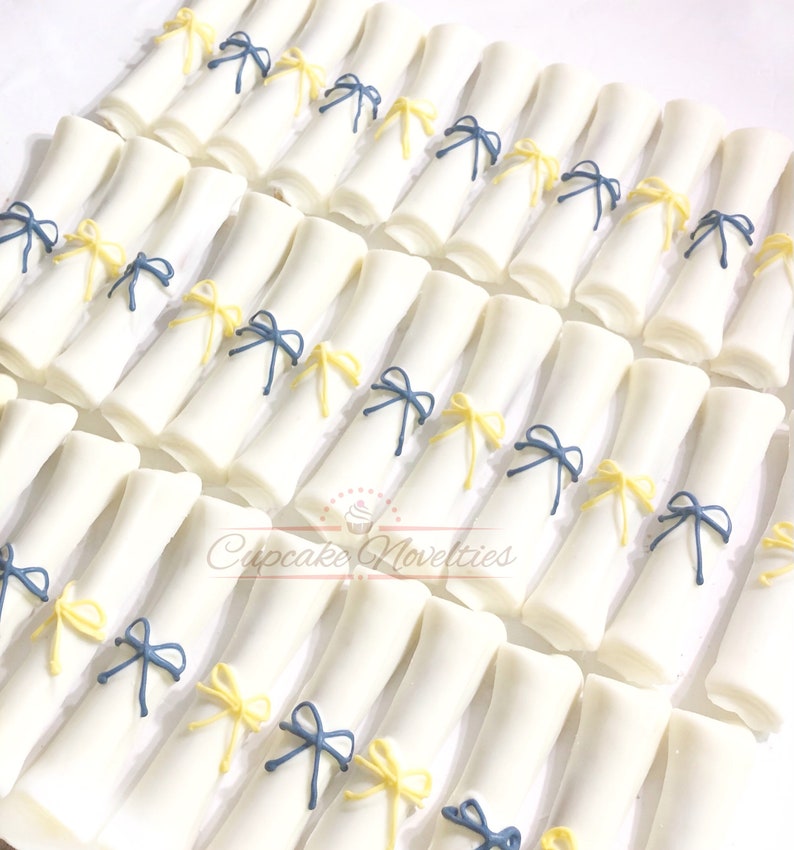 May include: A collection of white fondant graduation caps decorated with yellow and blue ribbon bows. The caps are arranged in rows on a white surface.
