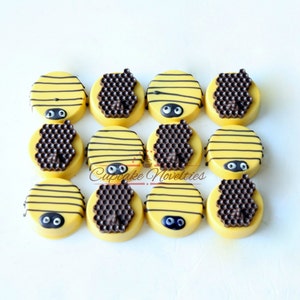 May include: Twelve yellow-frosted cookies decorated to resemble bees. Each cookie has black stripes, a chocolate honeycomb, and two black candy eyes. The cookies are arranged on a white surface. The text "Cupcake Novelties" is visible.