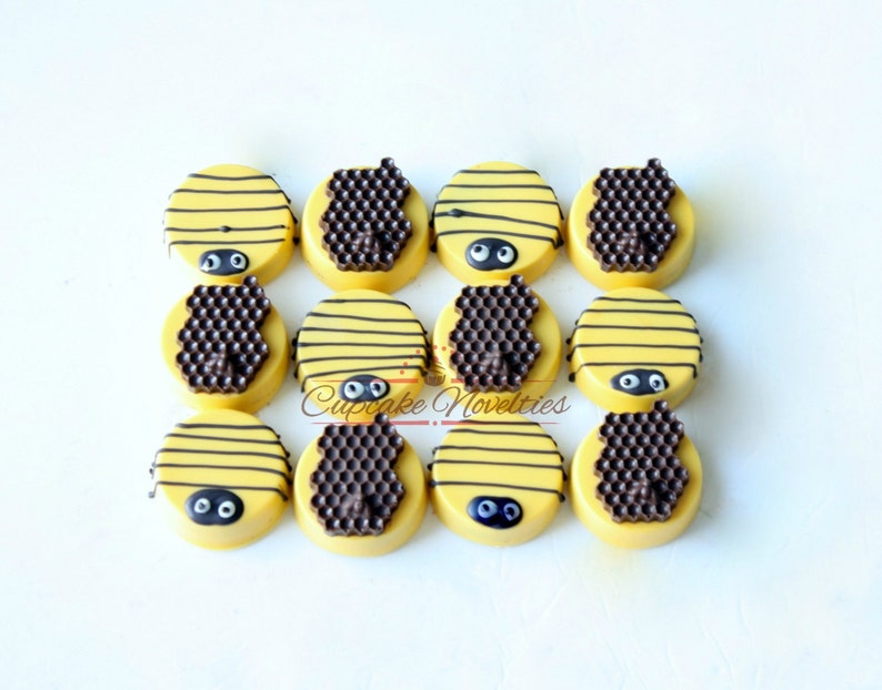 May include: Twelve yellow-frosted cookies decorated to resemble bees. Each cookie has black stripes, a chocolate honeycomb, and two black candy eyes. The cookies are arranged on a white surface. The text "Cupcake Novelties" is visible.