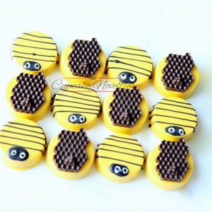 May include: A dozen yellow-frosted Oreo cookies decorated to resemble bees. Each cookie has black stripes, a chocolate honeycomb, and two black and white candy eyes. The cookies are arranged on a white surface.