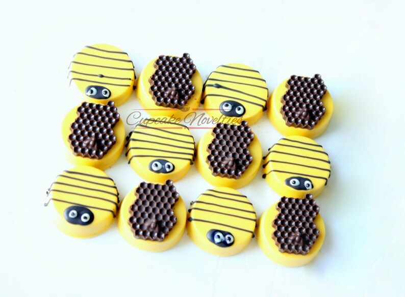 May include: A dozen yellow-frosted Oreo cookies decorated to resemble bees. Each cookie has black stripes, a chocolate honeycomb, and two black and white candy eyes. The cookies are arranged on a white surface.