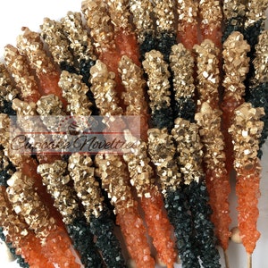 May include: A collection of gold, orange, and green rock candy sticks on wooden sticks. The candy is arranged in a fan shape.