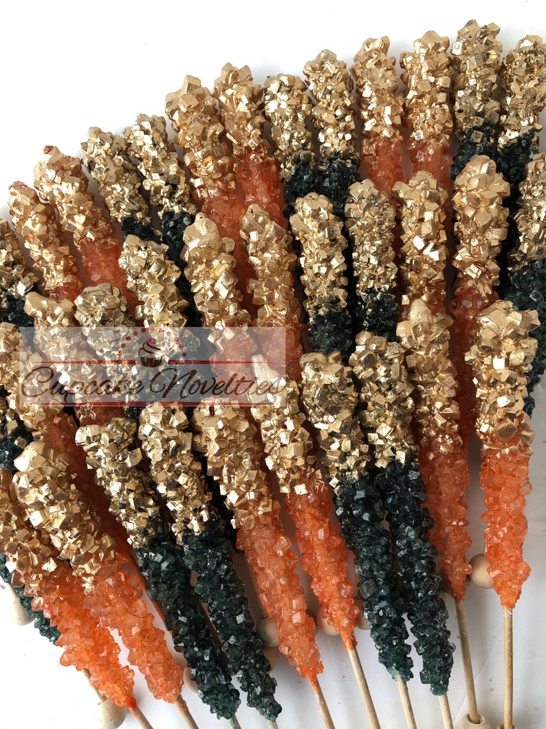 May include: A collection of gold, orange, and green rock candy sticks on wooden sticks. The candy is arranged in a fan shape.