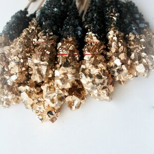 May include: A bunch of black and gold rock candy sticks on white sticks. The candy is arranged in a fan shape.