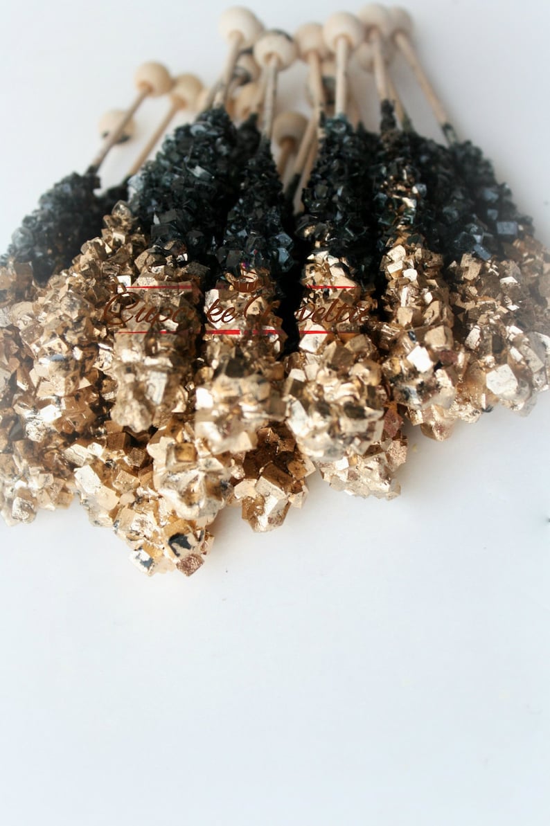 May include: A bunch of black and gold rock candy sticks on white sticks. The candy is arranged in a fan shape.