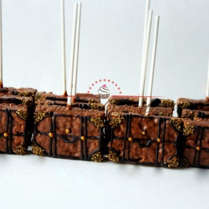 May include: A row of chocolate-covered rice crispy treats shaped like treasure chests, decorated with gold sprinkles and chocolate accents, on white sticks.