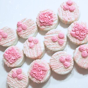 May include: A dozen white chocolate covered cookies decorated with pink flowers and white sprinkles.