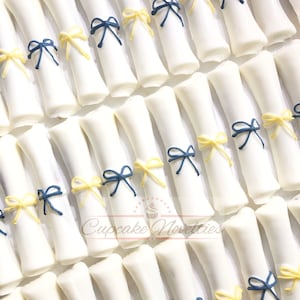 May include: White chocolate graduation cap favors decorated with blue and yellow bows. The favors are arranged in rows on a white surface. Cupcake Novelties is printed on the surface.