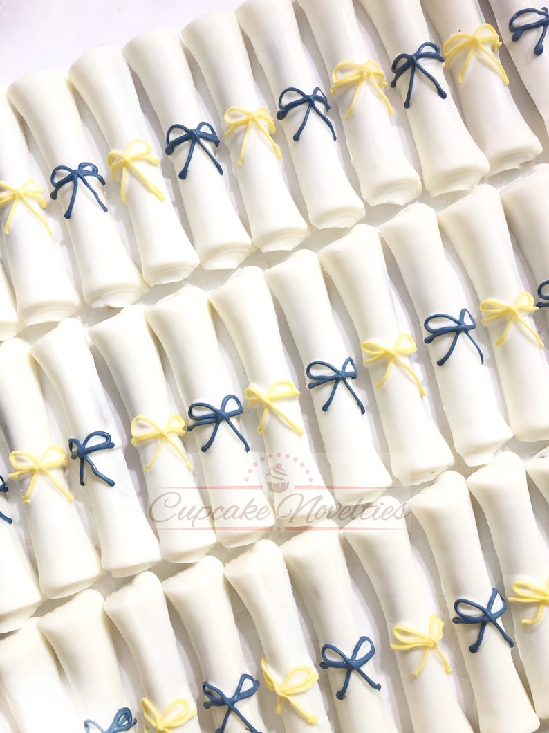 May include: White chocolate graduation cap favors decorated with blue and yellow bows. The favors are arranged in rows on a white surface. Cupcake Novelties is printed on the surface.