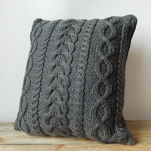 May include: A square, gray knitted pillow with a cable knit design. The pillow features vertical cable patterns of varying widths and textures, creating a cozy and textured appearance. The pillow is made of a soft, knitted material.