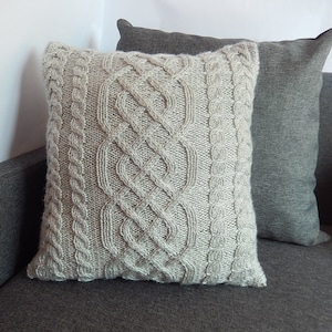 May include: A beige knitted throw pillow with a cable knit pattern. The pillow is on a gray couch.