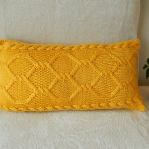 Op de afbeelding: Een geel gebreid kussen met een kabelbreipatroon. Het kussen is rechthoekig en heeft een zachte, pluche textuur.