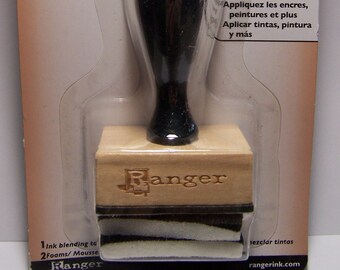 Può includere: Uno strumento di miscelazione dell'inchiostro in legno marrone con un manico nero e un cuscinetto di schiuma bianco e nero. Lo strumento è etichettato "Ranger" e "Ink blending tool, Applicateur d'encre, Herramiento para mezclar tintas".