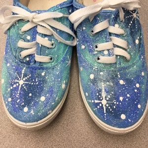 May include: A pair of blue canvas sneakers with white paint splatters and a galaxy design. The inside of the shoes are white with the words "Faded Glory" printed in pink.