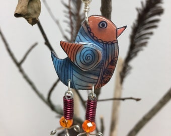 May include: A blue and orange bird charm with silver wire legs and orange beads. The bird has a swirl pattern on its body and is hanging from a silver wire.