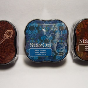Könnte beinhalten: Drei StazOn-Stempelkissen in dekorativen Dosen. Eines ist braun mit einem Löffeldesign und dem Text "Spiced Chai". Das zweite ist blau mit einer Meerjungfrau und dem Text "Blue Hawaii". Das dritte ist braun mit einem Uhrendesign und dem Text "Ganache".