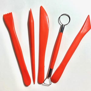 May include: A set of five bright orange sculpting tools for clay work. The collection includes a serrated edge tool, a pointed stylus, a flat blade, a wire loop tool with a metal head, and a flat angled spatula tool, all featuring smooth plastic handles.