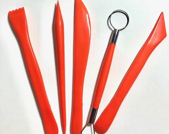 Può includere: Un set di cinque strumenti per scultura di colore arancione brillante per argilla. La collezione include uno strumento con bordo seghettato, uno stilo appuntito, una lama piatta, un utensile a filo con testa in metallo e una spatola piatta angolata.