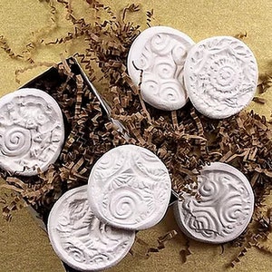 May include: A collection of six white, round, decorative stones with various embossed designs, including swirls, leaves, and abstract patterns. The stones are arranged in a black box filled with brown paper shreds, set against a yellow-tan background.