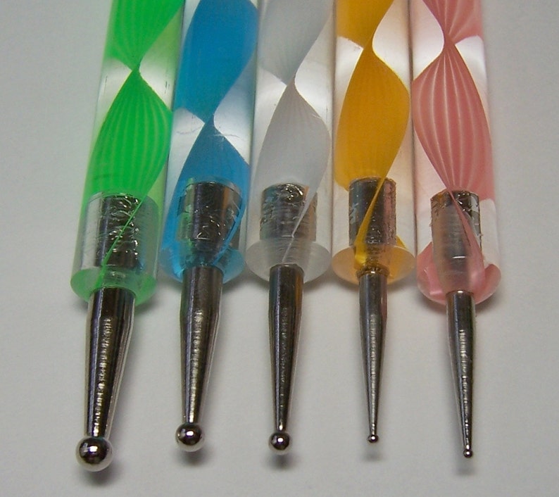 Pu&ograve; includere: Cinque strumenti per nail art colorati con manici di diversi colori e punte metalliche. I manici sono verde, blu, bianco, giallo e rosa con un motivo a spirale. Le punte metalliche sono rotonde e appuntite.