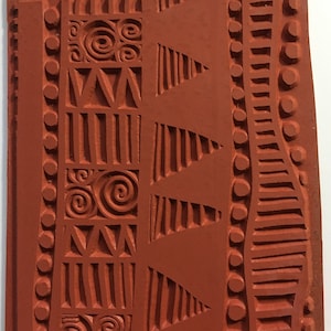 May include: A rectangular, reddish-brown rubber stamp with an abstract design. The design includes geometric shapes like triangles, vertical lines, and spirals, along with a series of circles and curved lines. The stamp is approximately 3 inches wide.
