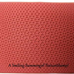 Puede incluir: Un sello de textura de goma roja con un patrón repetitivo de pequeños triángulos. El sello está diseñado para crear un efecto texturizado en papel u otras superficies. El texto "A Smiling Summergirl Texture Stamp!" está impreso en la parte inferior del sello.