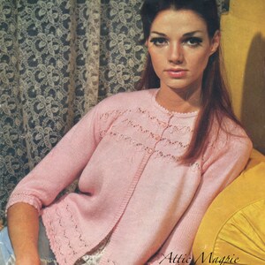 May include: A pink knitted cardigan with a delicate lace pattern around the neckline and cuffs. The cardigan has a button closure and is worn by a woman with long red hair.