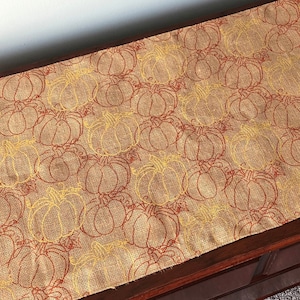 May include: A brown burlap table runner with a repeating pattern of yellow and orange pumpkins. The runner is on a dark wood surface.