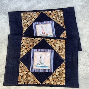 May include: Two blue denim placemats with a woodgrain pattern and a sailboat design in the center. The sailboats are white with blue and red accents.