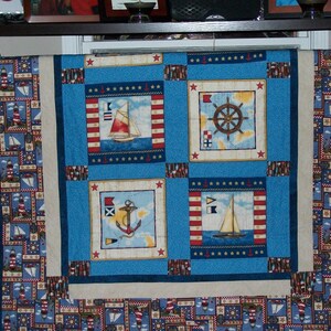 May include: A blue and white nautical themed quilt with a border of lighthouses and sailboats. The centre of the quilt features four squares with different nautical designs, including a ship's wheel, an anchor, and two sailboats.