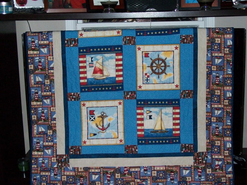 May include: A blue and white nautical themed quilt with a border of lighthouses and sailboats. The centre of the quilt features four squares with different nautical designs, including a ship's wheel, an anchor, and two sailboats.