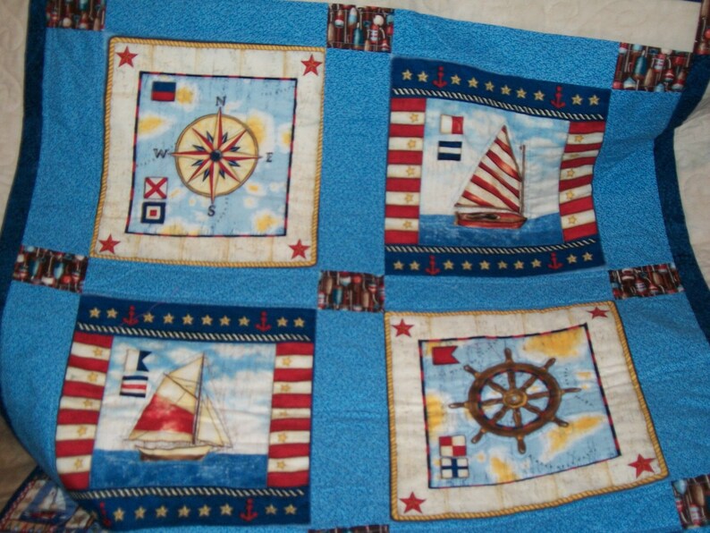 May include: A blue and white nautical themed quilt with four squares. Each square features a different nautical design: a compass, a sailboat, a ship's wheel, and a sailboat with a red, white and blue striped sail.