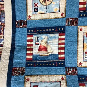May include: A blue and white nautical themed quilt featuring a compass, sailboat, and anchor designs. The quilt is made from fabric squares with a variety of patterns and colours.