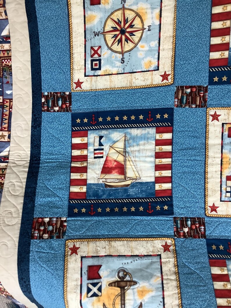 May include: A blue and white nautical themed quilt featuring a compass, sailboat, and anchor designs. The quilt is made from fabric squares with a variety of patterns and colours.