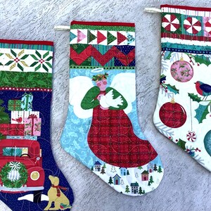 May include: Three quilted Christmas stockings with festive designs. One features a red truck with a Christmas tree and presents, another an angel, and the third has ornaments and a bird. Each has a patterned top.