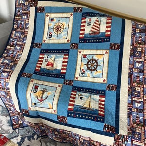 May include: A blue and white nautical themed quilt featuring a variety of images including a compass, a sailboat, an anchor, and a ship's wheel. The quilt is made with a variety of fabrics and has a white border.