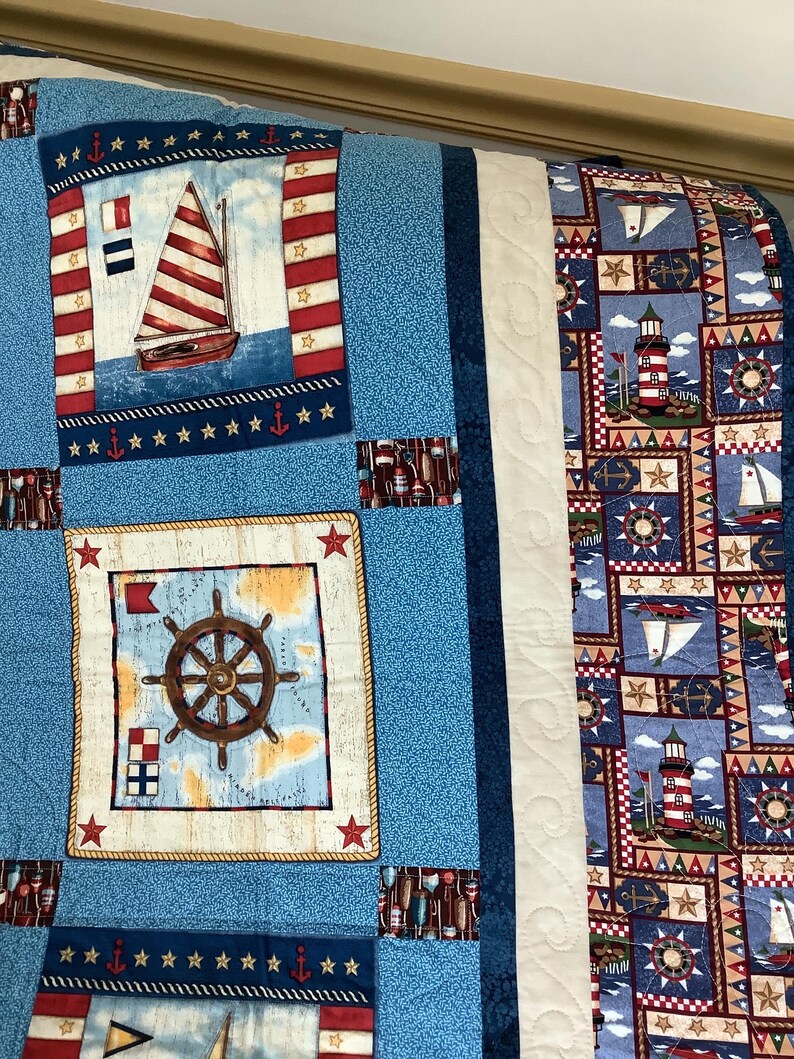 May include: A blue and white nautical themed quilt with a white border. The quilt features a sailboat, a lighthouse, a ship's wheel, and stars.