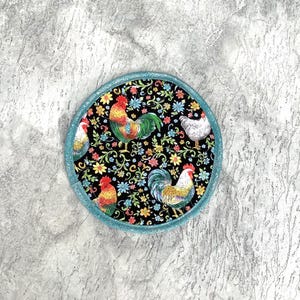 May include: Round coaster with a black background featuring colorful roosters and floral patterns. The coaster has a teal border. The roosters are red, yellow, green, and white. The flowers are yellow, blue, and pink.