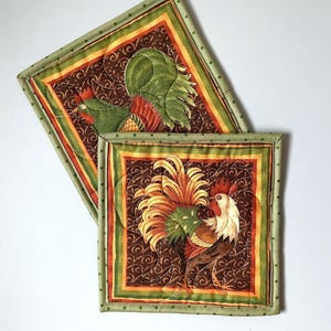 May include: Two square quilted pot holders featuring rooster designs. The pot holders have a brown background with a repeating swirl pattern. The roosters are in shades of green, red, orange, and white. The borders are green with a dotted pattern.