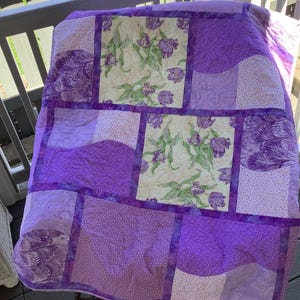 May include: A handmade quilt featuring a patchwork design in shades of purple and white. The quilt incorporates floral patterns and solid fabrics, with a border of purple fabric. The quilt is displayed outdoors.
