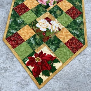 May include: A quilted table runner with a Christmas theme. It features a patchwork design with green, red, gold, and white fabrics. The runner has a floral pattern with poinsettias, roses, and pine branches.