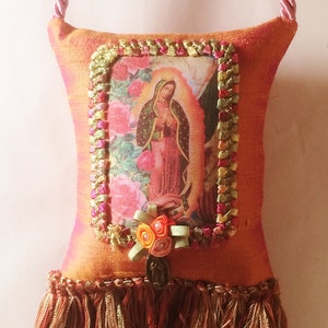 May include: Orange fabric pillow with a fringed bottom. The pillow features a framed image of the Virgin Mary with a floral background. The frame is decorated with colorful ribbons and a small flower embellishment.