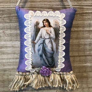 May include: A purple satin pillow with a white lace trim and a picture of an angel with wings. The pillow has a fringe trim and a purple flower.