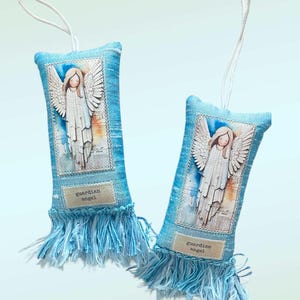 May include: Two blue fabric ornaments with a printed image of an angel with outstretched wings. The ornaments have a fringe at the bottom and a tag that reads "guardian angel". Each ornament has a white string for hanging.