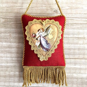 May include: Red decorative pillow with a gold heart-shaped design featuring an angel illustration. The pillow has a gold lace trim, a gold rope handle, and a gold fringe at the bottom.